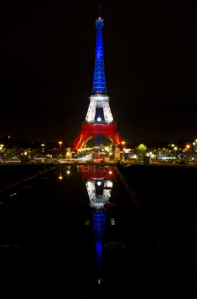 Night view of the tour Eiffel illuminated with bleu blanc rouge lights, reflecting the French flag colors. The tower's reflection is visible in a calm water surface below. Surrounding city lights and dark sky frame the iconic structure.