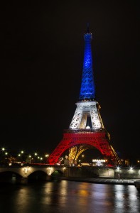 The tour Eiffel illuminated with bleu, blanc, rouge lights, representing the colors of the French flag, against a black night sky. The reflection of the tower and lights can be seen on the water below.