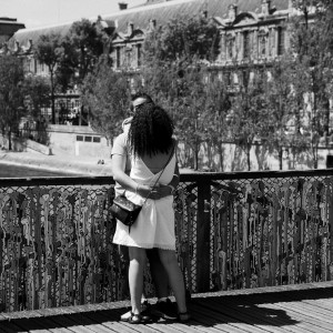 A couple amoureux embraces and kisses on the pont des Arts, surrounded by decorative metal panels, with historic buildings and trees in the background, captured in classic black and white.