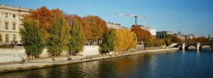 A riverside scene on the Quai de Seine in Paris, with people relaxing along the Seine, autumn trees displaying colorful automne foliage, historic buildings, a bridge, and a construction crane under a clear blue sky.