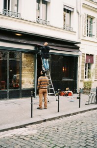 A person stands on a ladder working above the entrance of a building, while another person stands below, watching. The scene is on a cobblestone street in front of a shopfront with large windows.