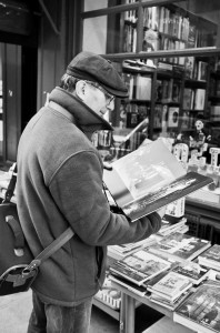 A man wearing a flat cap, glasses, and a jacket stands by a table of books outside a shop in Paris, looking at a large open book. Shelves filled with books are visible through the window behind him, suggesting a quiet consultation at minuit.