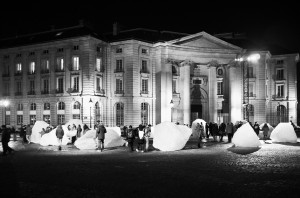 A black and white photo captures a night scene outside a grand building with columns and multiple windows. People are gathered around large ice formations, part of an "Ice Watch" public art installation, spread across the cobblestone plaza in front of the building, creating a dynamic spectacle.