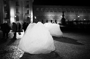 A black and white photo captures large ice blocks placed on an outdoor cobblestone plaza at night. Groups of people gather around, engaging with the art installation by the Ice Watch brand. Historic buildings in the background are illuminated by streetlights.