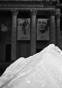 Black and white photo showing a close-up of a large stone with a historic building in the background. The building, blurred by time like an ancient watch, has tall columns and two large banners displaying portraits of individuals. The stone dominates the foreground, grounding the scene in permanence.