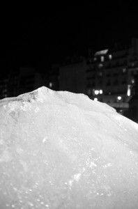 A large, glistening ice formation is prominently displayed in the foreground under a dark night sky. In the distant background, blurry city buildings and street lights create a contrasting backdrop. An abandoned Ice Watch lies buried within the ice, adding an intriguing layer to this black-and-white scene.
