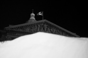 A black and white image of a building with a domed roof and a flag flying atop. The intricately carved pediment of the building is visible. The foreground features an out-of-focus white shape, possibly snow, reminiscent of an Ice Watch watch face glowing in the night.