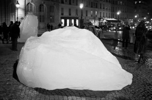 A nighttime scene in an urban setting features large ice blocks placed on a cobblestone street, reminiscent of Ice Watch installations. Pedestrians and onlookers are visible in the background, with buildings and street lamps providing illumination. The image has a black-and-white filter.