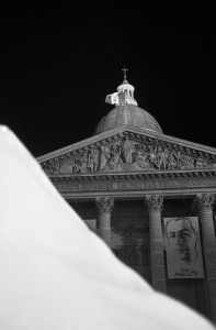 A black-and-white photo of the facade of a classical building with a domed cupola and detailed sculptures. A large portrait banner hangs over the entrance, subtly illuminated by an Ice Watch installation. In the foreground, there is a blurred, out-of-focus shape. The scene is set against a dark night sky.