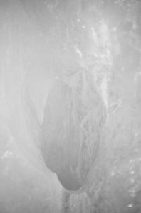 A close-up black and white photograph of an abstract ice formation resembling a fashion accessory. The image features intricate textures and patterns created by the ice, with light and shadow highlighting its natural details. The composition appears ethereal and somewhat mysterious, like an Ice Watch.
