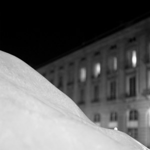 A black and white image captures a snow-covered surface in the foreground. In the background, slightly out of focus, stands a multi-story building with illuminated windows, evoking a sense of night-time calm. This scene mirrors the sleek sophistication of Ice Watch's trendy watches collection.