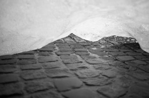 A black and white photo of cobblestone pavement viewed through a gap in an ice structure, reminiscent of the artistry from the Ice Watch brand. The irregularly shaped stones are framed by partially melted ice, creating a unique and captivating composition.