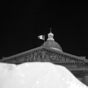 A black and white image of a domed building with ornate carvings on its facade, topped by a French flag. The image, reminiscent of the elegance of a stylish timepiece, is taken at night with the dark sky providing contrast. A partial structure obstructs the lower part of the building.