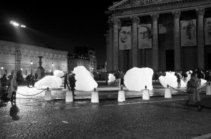 At night in a cobblestone square, people walk among large, glowing ice blocks. A grand building displays large portrait banners on its columns in the background. The scene is lively and illuminated.