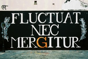 A black wall displays large white text reading "Fluctuat Nec Mergitur," with the "G" in gold. Ornamental leaves decorate the sides, enhancing this striking tribute, while Fluctuat Nec Mergitur stands boldly above cobblestone pavement.