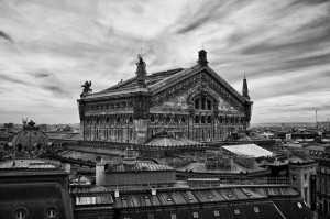 A black and white photo of the ornate Opéra Garnier in Paris, featuring detailed sculptures and architectural elements, with dramatic cloudy skies above and city rooftops in the foreground.