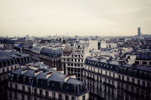 A cityscape view of Paris in hiver with classic Haussmann-style buildings, gray rooftops, and chimneys under a cloudy sky; the Montparnasse Tower is visible in the distance.