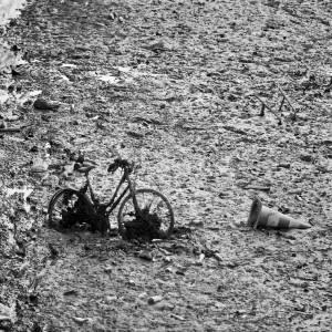 A black and white photo showing a muddy, barren landscape near Canal Saint-Martin with a partially buried bicycle in the mud. A tipped-over traffic cone lies nearby on the right side. The ground is strewn with debris and tire tracks, remnants of a recent vidange process in Paris.