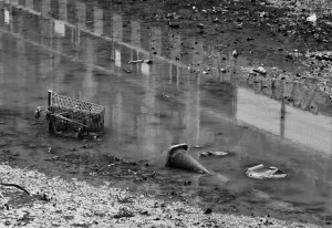A black and white image depicts a muddy, litter-strewn puddle near Canal Saint-Martin. The foreground features a tipped-over shopping cart and a traffic cone partially submerged in the water. Various pieces of trash and debris are scattered around, creating a scene reminiscent of Vidange. Reflective surfaces indicate nearby buildings.
