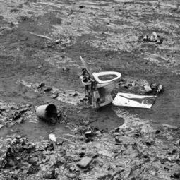 A discarded toilet, a broken bucket, and scattered debris lie in a muddy, litter-strewn environment. The black and white image captures the scene of environmental neglect and pollution around Canal Saint-Martin after a vidange.