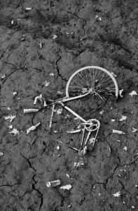 A black-and-white photo of a broken and abandoned bicycle lying on dry, cracked ground. The bicycle frame is bent and missing several parts. Empty cans and fallen leaves are scattered around the bike, reminiscent of scenes during the Vidange du Canal Saint-Martin. The scene conveys neglect and desolation.