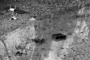 A black and white photograph of a muddy, shallow water body resembling the Canal Saint-Martin with various discarded items scattered around, including a chair and a rectangular container. The scene appears desolate and abandoned, akin to the vidange process, with reflections of nearby objects visible in the water.