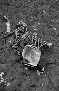 A black and white image of a broken and overturned shopping cart lying on cracked, dried mud evokes scenes of neglect. Debris and litter scattered around the cart further highlight this abandoned spot near Canal Saint-Martin, reminiscent of a vidange after the water has drained away.