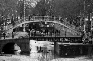 A black and white photo of Canal Saint-Martin shows a bridge crowded with people over a river that appears to be partially drained. The vidange reveals the riverbed littered with debris, ducks, and some bicycles. Trees without leaves line the riverbanks, and buildings are visible in the background.