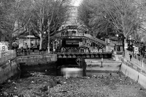 A black and white image shows the Vidange of Canal Saint-Martin in a city, mostly drained with some water at the bottom. A bridge with intricate metalwork crosses overhead, and trees line both sides of the canal. People walk on the bridge and around the canal, and buildings are visible in the background.