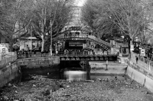 A black and white image shows the Vidange of Canal Saint-Martin in a city, mostly drained with some water at the bottom. A bridge with intricate metalwork crosses overhead, and trees line both sides of the canal. People walk on the bridge and around the canal, and buildings are visible in the background.