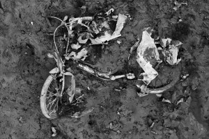 A black and white image showing a rusted, decayed bicycle partially buried in mud and debris near Canal Saint-Martin. The frame is bent and broken, with once-functional parts obscured by layers of dirt and fallen leaves, indicating long-term neglect and abandonment reminiscent of a vidange.