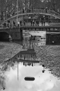 A black-and-white photo of the Canal Saint-Martin during its vidange shows a drained canal with a narrow stream of water and scattered debris. Many people stand on two pedestrian bridges above, while bare trees are visible in the background. A tire is partially submerged in the water.
