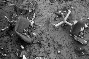 A black and white image showing broken and discarded office chairs submerged in muddy water, reminiscent of a vidange cleanup. The ground along what appears to be Canal Saint-Martin is littered with leaves, debris, and a crushed aluminum can. The scene suggests abandonment and neglect.