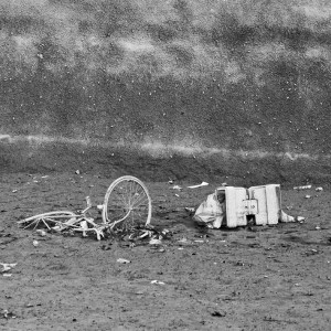 A monochromatic image showing a derelict bicycle and a broken television set half-buried in the mud, surrounded by scattered debris. The background comprises a textured, sloping wall, adding to the desolate atmosphere reminiscent of Canal Saint-Martin post-vidange.