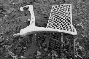 A black and white image of an overturned shopping cart lodged in muddy ground near Canal Saint-Martin, surrounded by fallen leaves. The cart's wheels are visible, and the metal grid structure is partially buried in the mud, creating a desolate, abandoned scene reminiscent of a Vidange.
