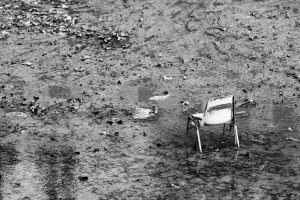 A desolate scene shows a single, worn chair half-submerged in a muddy, leaf-strewn area. The black-and-white photo captures an abandoned and forlorn atmosphere reminiscent of the Canal Saint-Martin vidange, emphasizing the solitary chair amidst the scattered debris.