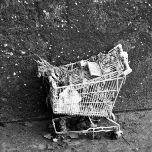 A black and white image of a rusty, overturned shopping cart leaning against a textured wall near Canal Saint-Martin. The cart, filled with dry leaves and debris, appears weather-beaten and abandoned. The ground around it is cluttered with fallen leaves, evoking a sense of vidange.