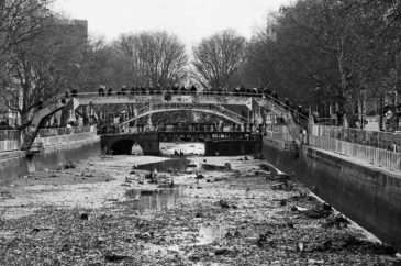 A black and white photo depicts the Canal Saint-Martin with significantly low water levels, revealing the rocky and muddy canal bed during its vidange. Two arched bridges span the canal, and numerous people are standing and observing on the bridges. Leafless trees line the sides of the canal.