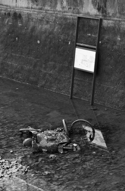 A black and white photo shows an abandoned bicycle partially submerged in the shallow, muddy area of Canal Saint-Martin. A concrete wall with a faded informational sign on a metal stand is in the background, suggesting recent vidange. The scene appears neglected and weathered.