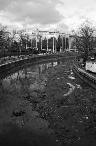 Black and white image of the Canal Saint-Martin during its vidange, with low water levels revealing mud and debris. Leafless trees line both sides of the canal, while buildings are visible in the background under a cloudy sky.