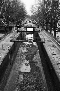 Black and white photo of an empty Canal Saint-Martin with exposed mud, surrounded by bare trees. A group of people stands on a bridge above, with a construction sign that reads "DODIN" visible halfway down the canal. The background is slightly foggy and overcast, capturing the essence of the vidange process.