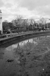 A grayscale photo of an urban canal with low water levels, exposing a muddy bed. Leafless trees line the Canal Saint-Martin, reflecting in the shallow water. Buildings and pedestrian walkways flank the canal, and the scene is overcast due to a recent vidange, giving a gloomy atmosphere.