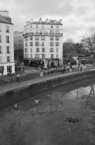 A black and white photo depicts an empty waterway, reminiscent of Canal Saint-Martin, in a city setting. In the background, several multi-story buildings line the street, with people walking along the sidewalk. The sky is overcast, and some trees are visible on the right side of the image.