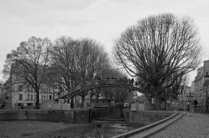 A black-and-white photo depicts a wooden arched bridge spanning over Canal Saint-Martin in an urban area. Leafless trees line both sides, and buildings are visible in the background. A group of people is walking across the bridge, with cobblestone streets nearby during the vidange season.