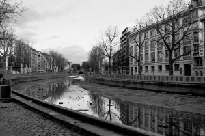 A black-and-white photograph of the Canal Saint-Martin with minimal water, possibly during its vidange, runs between rows of buildings on both sides. Leafless trees line the canal, reflecting in the shallow water. The sky is overcast, adding to the stark, somber mood of the scene.