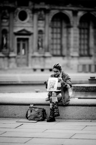 Anna Meunier, wearing sunglasses, sits on a stone bench sketching or painting on a pad. Dressed in a jacket and knee-high boots beside her bag, she’s framed by an ornate, blurred building in this black-and-white image.