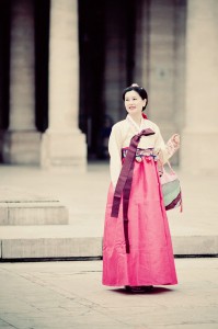 A woman stands outdoors wearing a traditional Korean hanbok with a Le bonbon rose pink skirt and cream top, holding a colorful bag and smiling. The background features tall stone columns and a paved area.