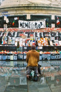 A person kneels in front of a memorial covered with candles, flowers, posters, and graffiti. French flags are visible among the tributes, alongside signs in French—including one boldly proclaiming "Même pas peur.