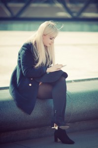 Magalie Delorme, a woman with long blonde hair, wearing a dark coat and black heeled boots, sits on a concrete bench outdoors, looking at her smartphone. It appears to be a cool, sunny day.