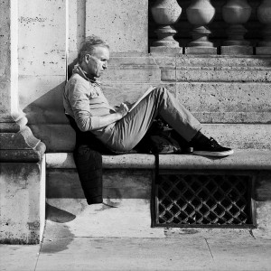 Lucien Robert sits on a stone bench outside, leaning against a wall and reading a book. He appears relaxed and focused, with one leg stretched out and the other bent as sunlight casts shadows across the scene.